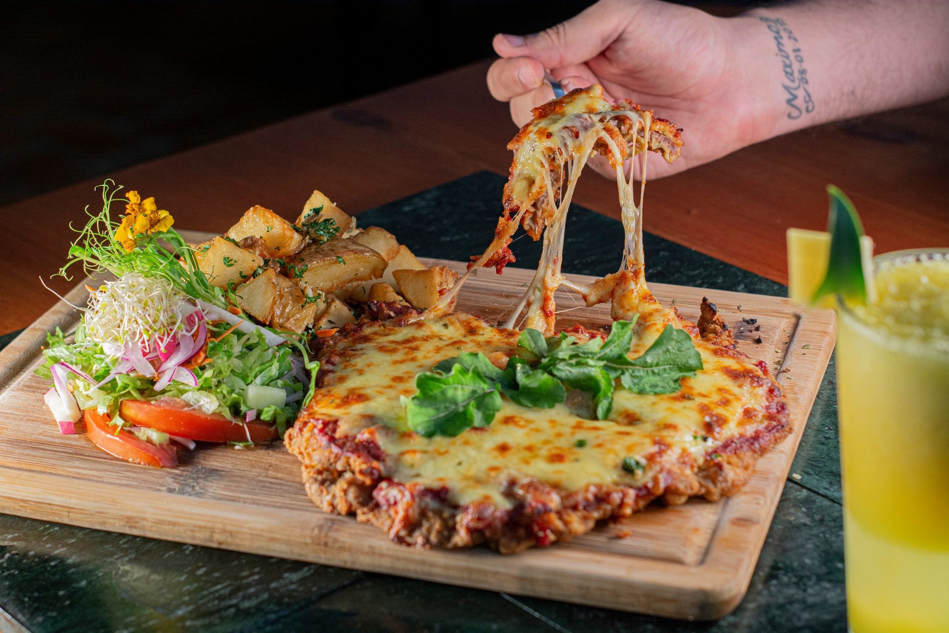 Tenedor levantando queso fundido de una milanesa con ensalada y papas